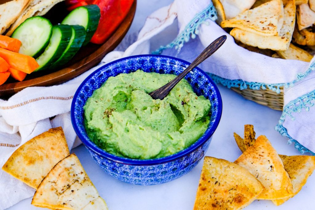 Spinach dip made with garbanzo beans in a blue serving dish with sliced vegetables and pita chips on the side. 