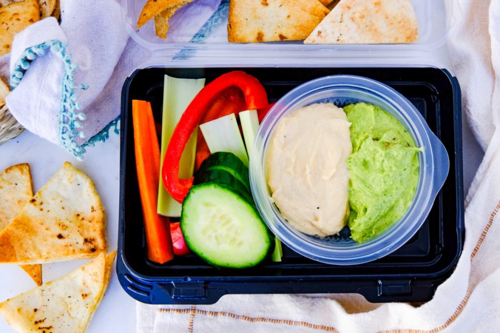 Hummus tray with vegetables in a plastic storage container with pita chips on the side. 