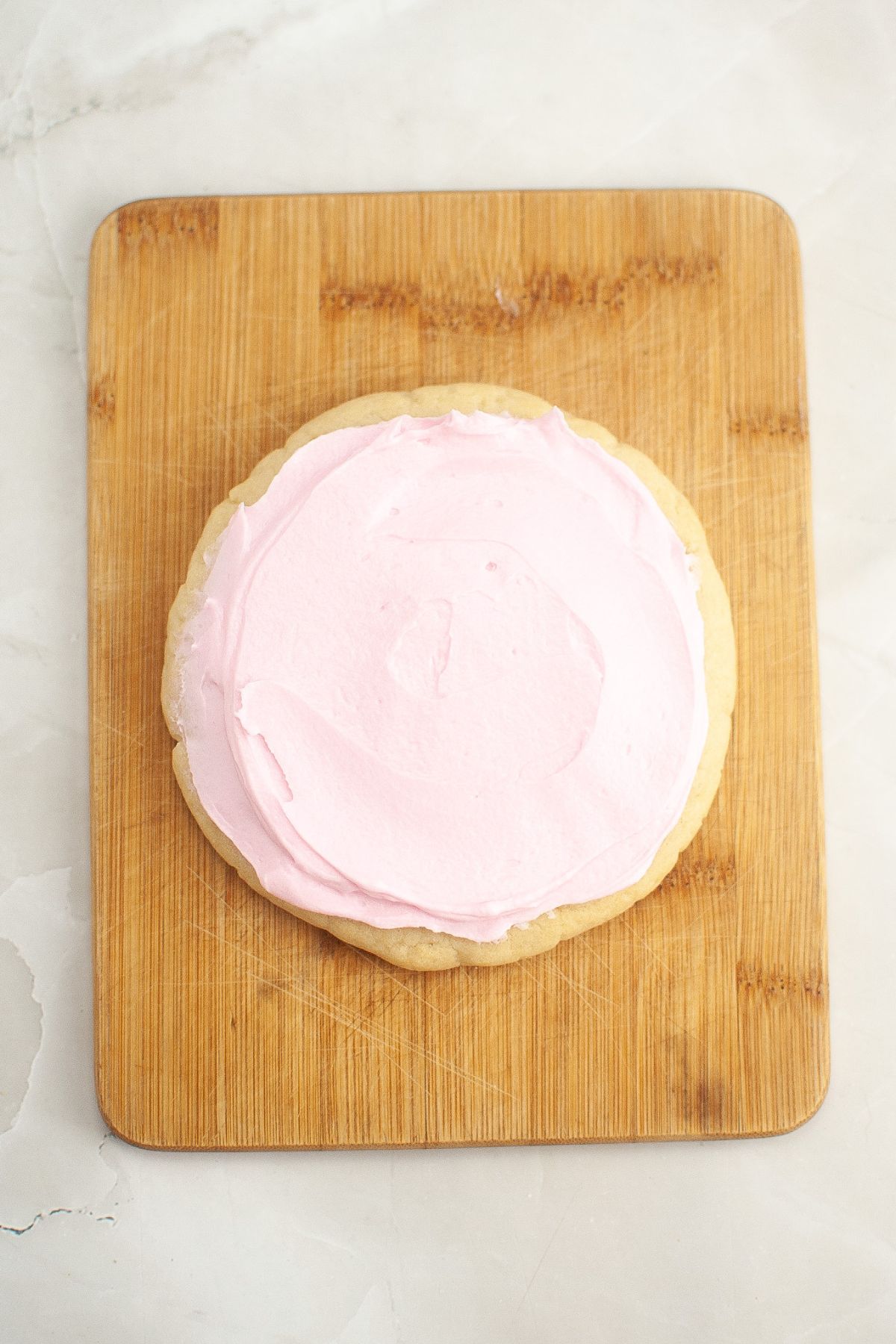 Pink frosting being spread on top of a cookie on a wooden cutting board.