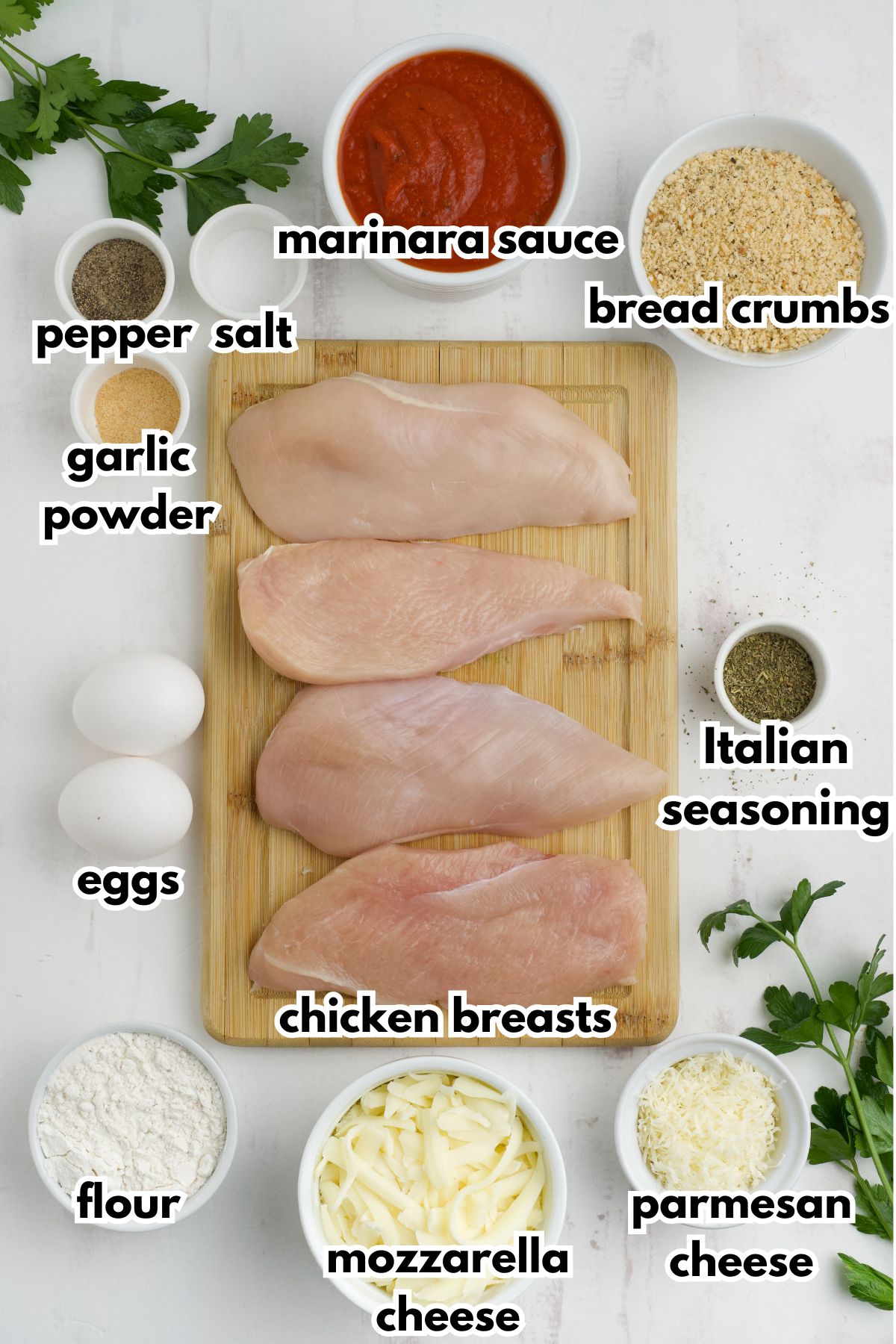 Bowls of pepper, salt, garlic powder, marinara sauce, bread crumbs, eggs, flour, chicken breasts, mozzarella cheese, parmesan cheese, and ltalian seaoning on a counter.
