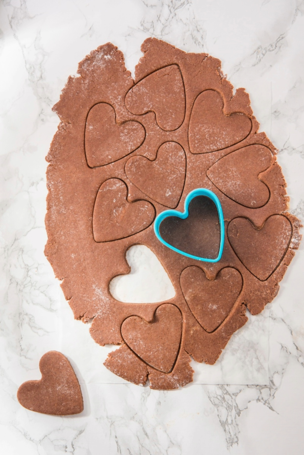 Cookie dough rolled on parchment paper with a blue heart cookie cutter pressed into heart shapes before baking.