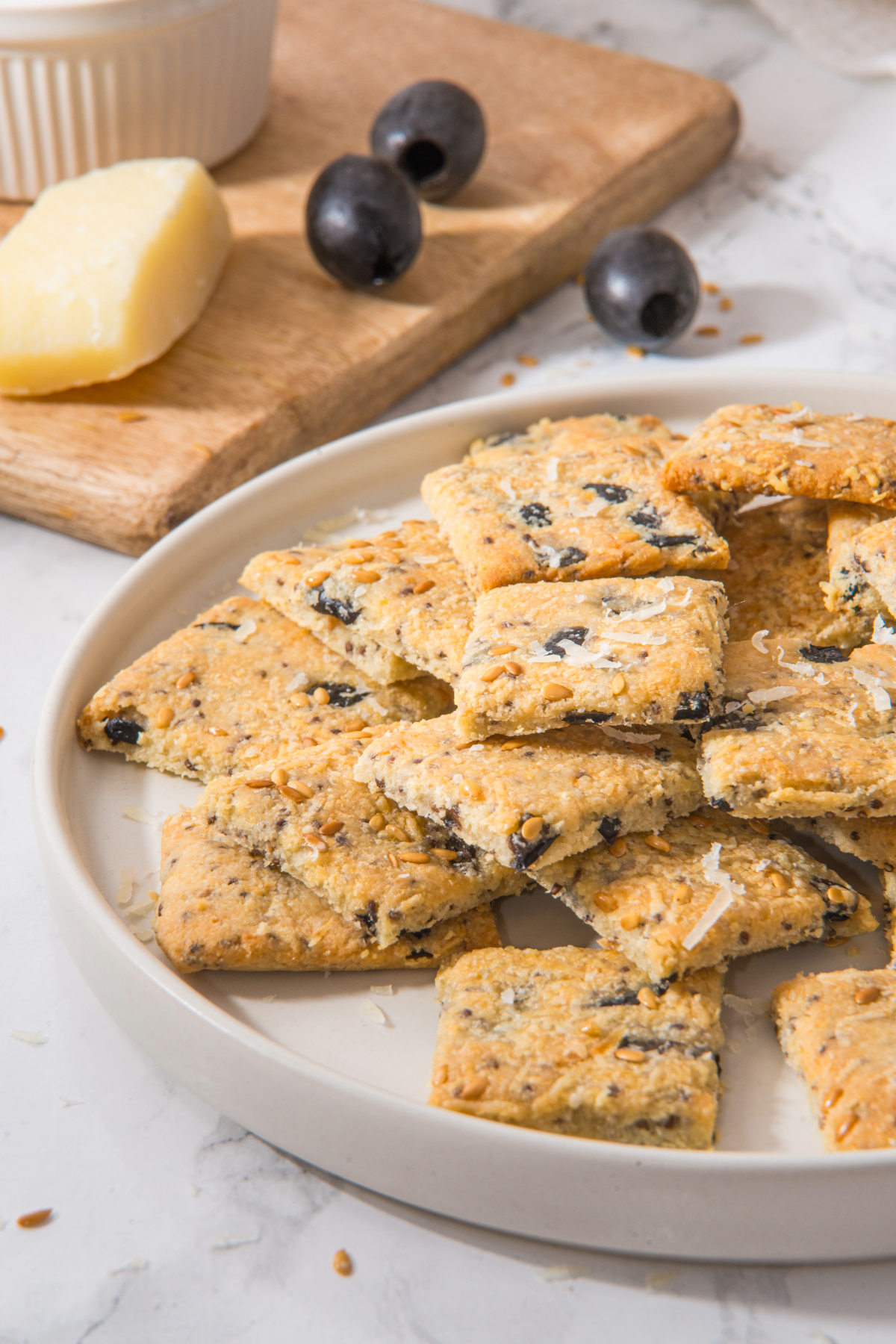 Small bite size keto crackers with chopped olives on a white serving plate.