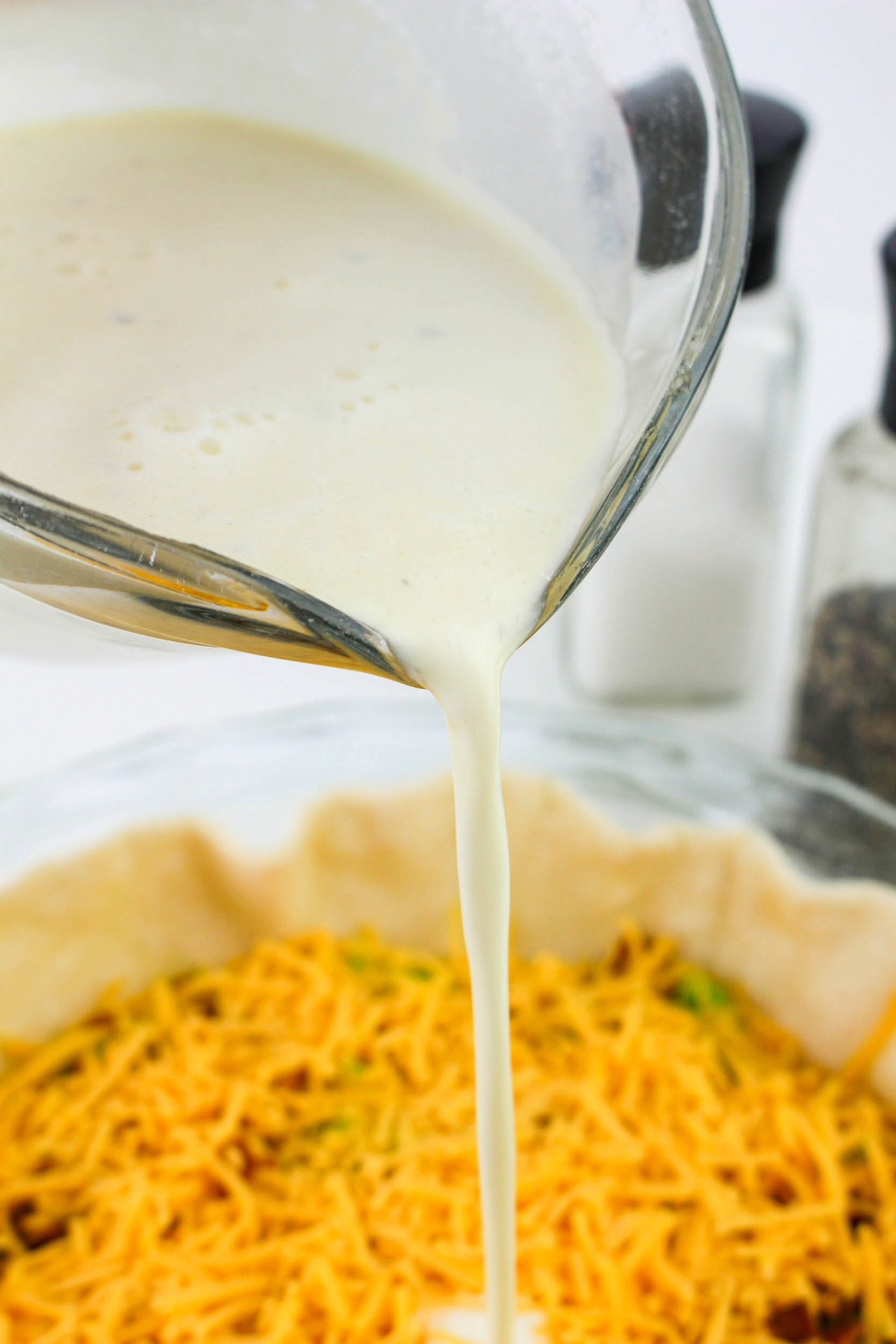 Milk, egg, and heavy cream mix pouring over quiche before baking.