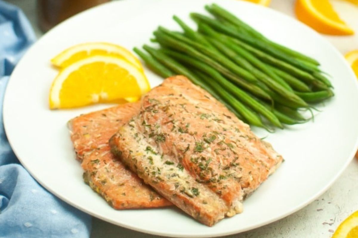 A white plate with two pieces of seasoned salmon fillets, a serving of green beans, and three slices of orange, placed on a blue cloth.
