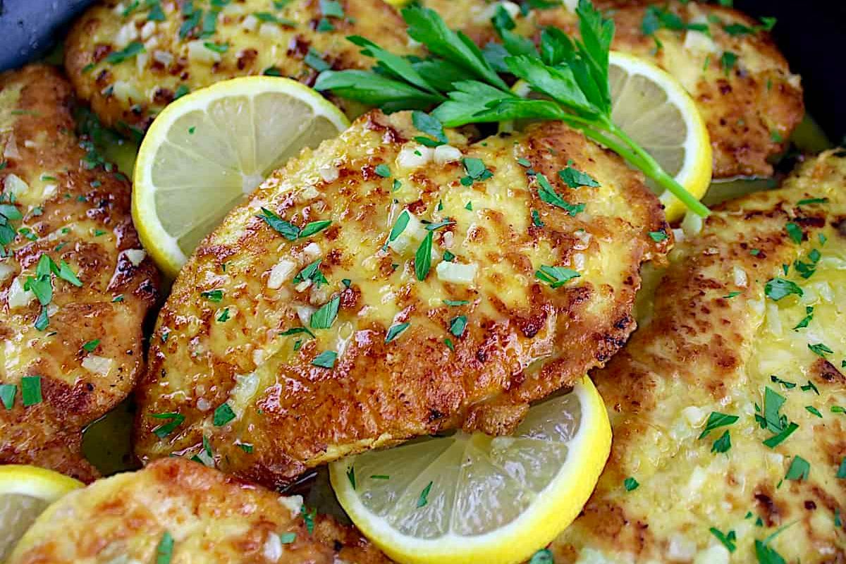 Close-up of a skillet with golden-brown chicken breasts garnished with chopped herbs and garlic. Lemon slices and a sprig of parsley are placed on top, enhancing the presentation. The dish appears freshly cooked and ready to serve.