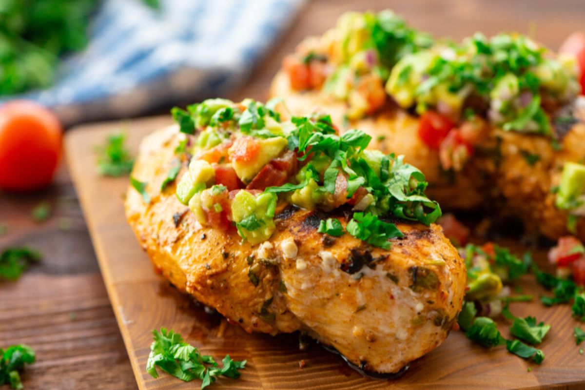 Grilled chicken breasts topped with a mixture of chopped tomatoes, avocado, and cilantro are placed on a wooden board. The dish is garnished with additional cilantro, with a blurred background featuring a blue cloth and a whole tomato.