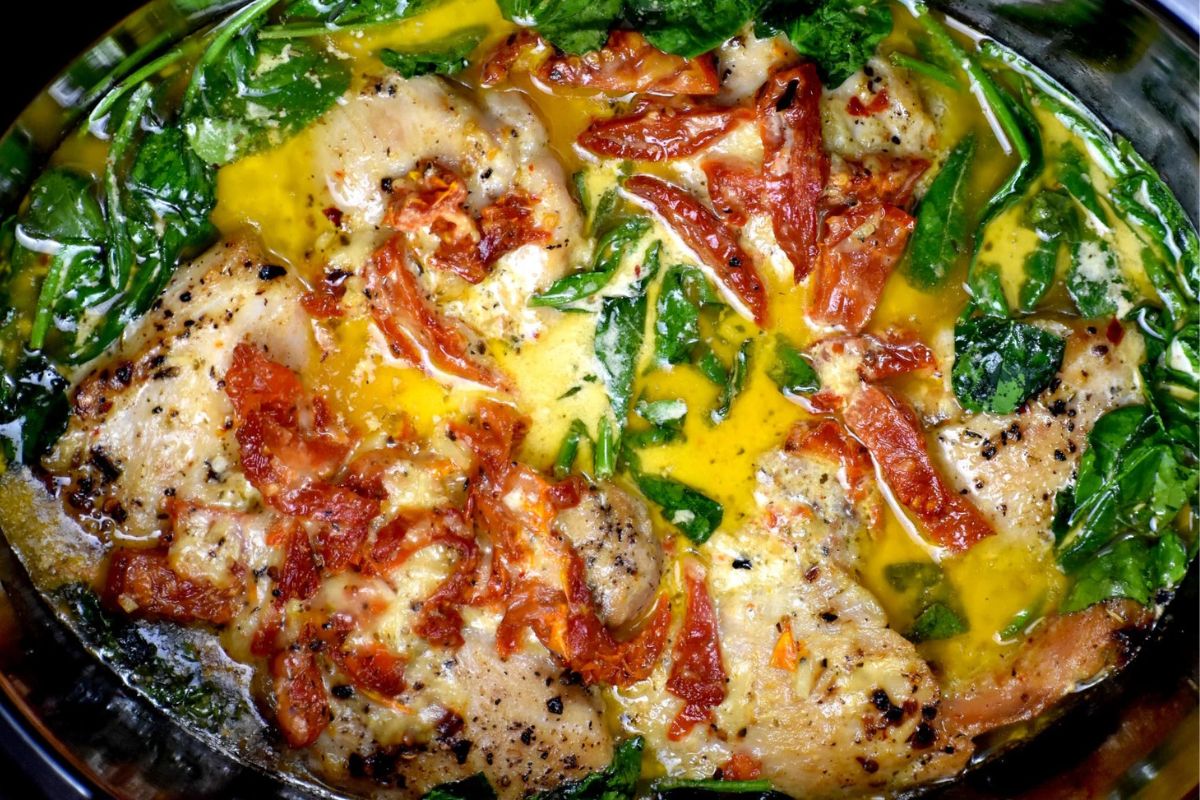 A slow cooker featuring seasoned chicken pieces, wilted spinach, and sun-dried tomatoes in a creamy, yellow sauce. The dish is garnished with black pepper and cooked in a metal pan.