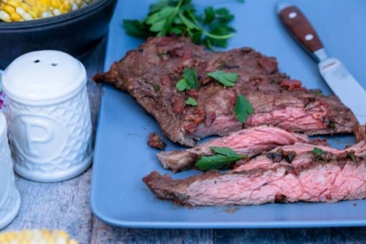 Sliced grilled steak garnished with parsley on a blue plate, alongside a knife. A white pepper shaker and a bowl of corn are partially visible in the background.