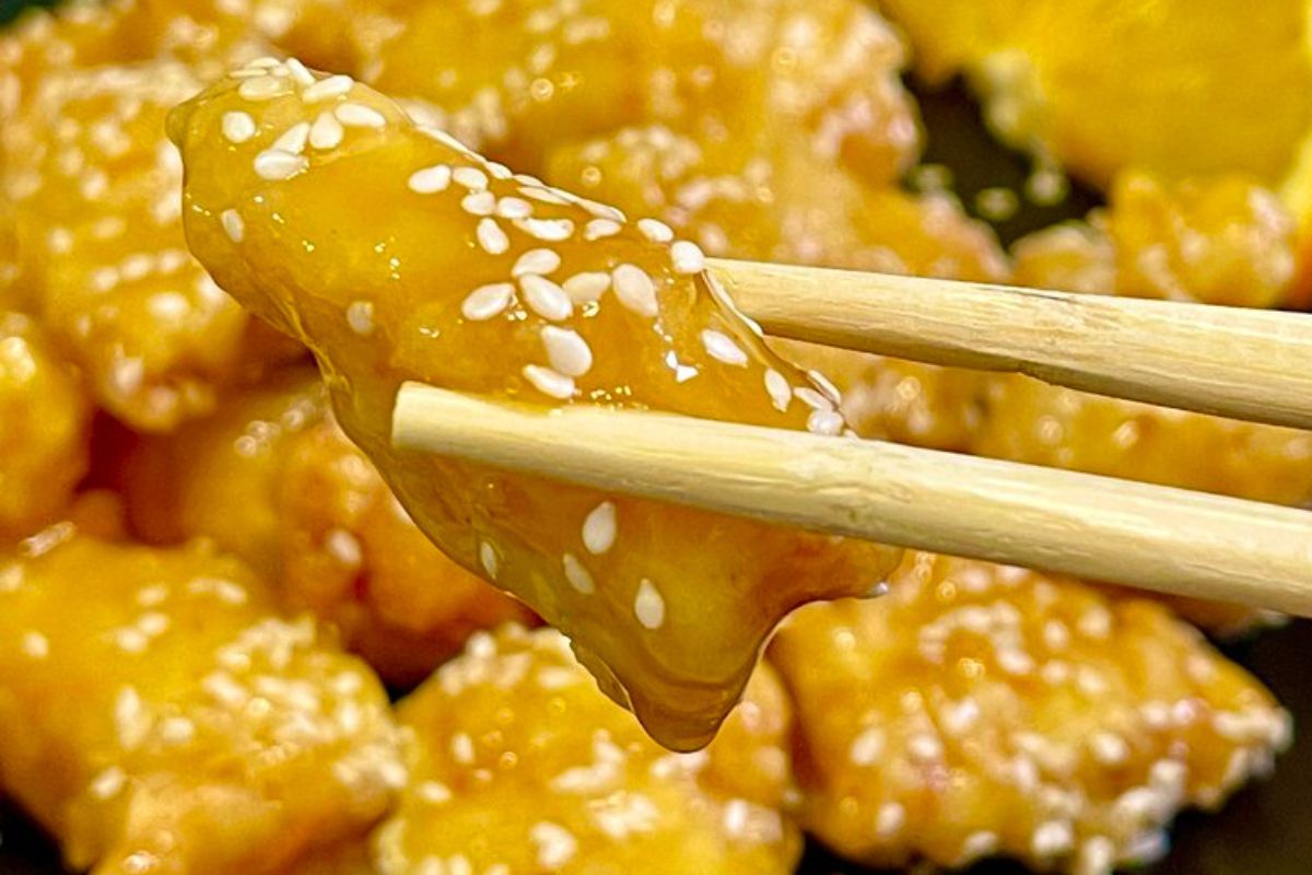A pair of chopsticks holds a piece of orange chicken covered in a sauce and sprinkled with sesame seeds. More pieces of orange chicken are visible in the blurred background.