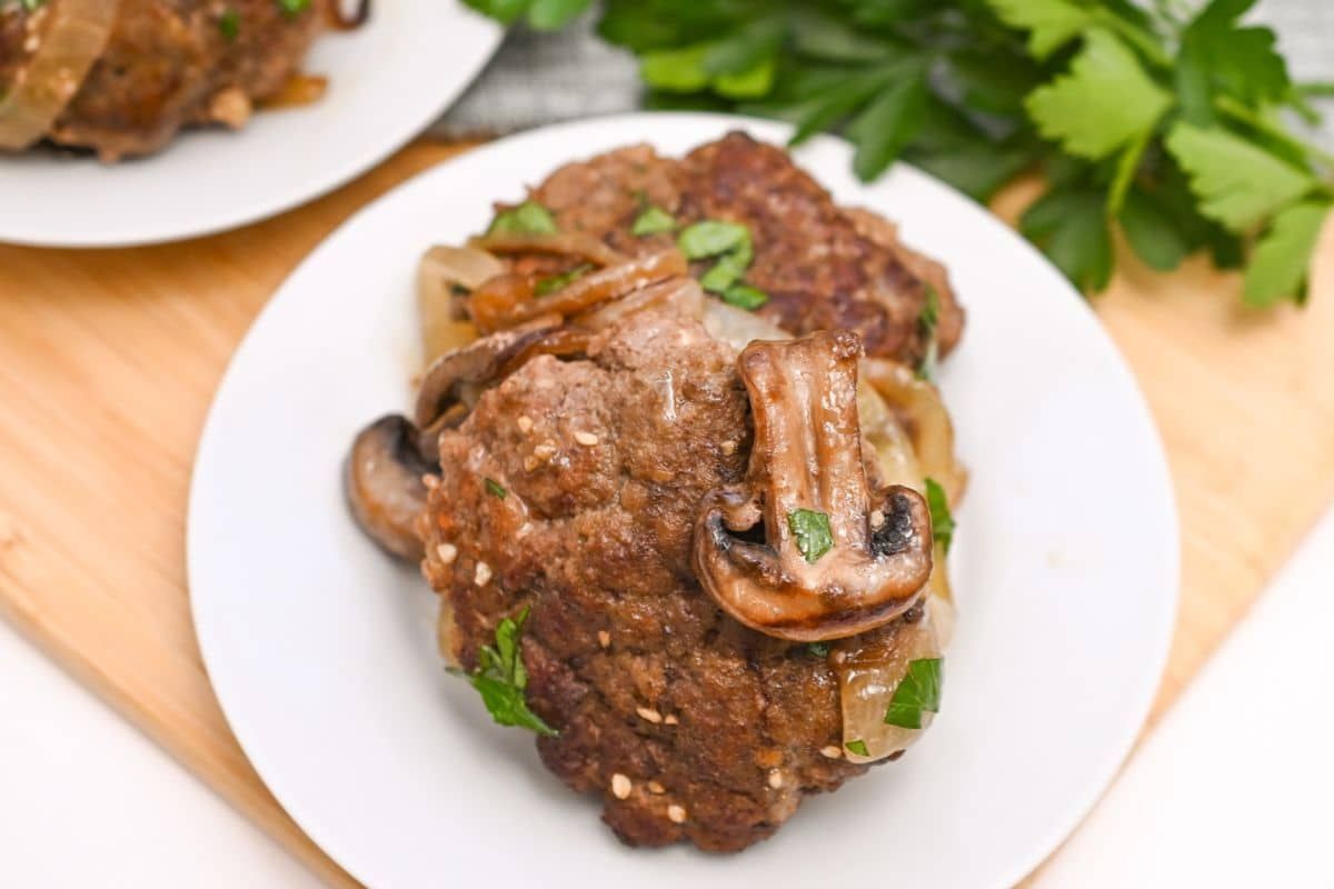 A plate with two cooked hamburger steaks topped with sautéed mushrooms, onions, and garnished with parsley. Fresh parsley leaves are visible in the background on a wooden surface.