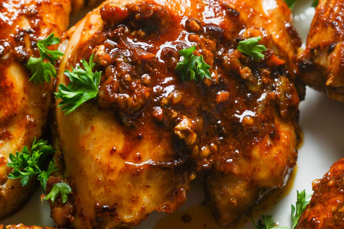 Close-up of a grilled chicken breast covered in a thick, dark sauce with visible herbs and spices. Fresh parsley leaves are scattered on top, adding a touch of green to the rich brown colors of the dish.