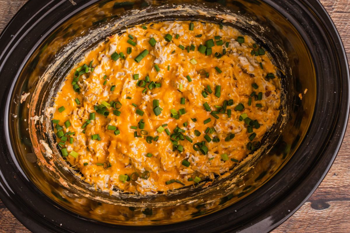 A slow cooker filled with a creamy mixture, topped with melted cheddar cheese and chopped green onions. The contents are a golden yellow with visible herbs, suggesting a savory dish. The slow cooker is set on a wooden surface.