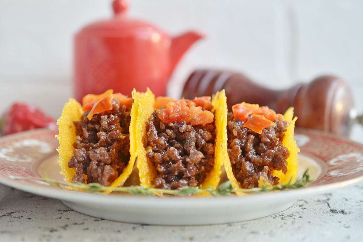 Three hard-shell tacos filled with seasoned ground beef and topped with diced tomatoes are arranged on a decorative plate. A red teapot and a pepper grinder are blurred in the background.