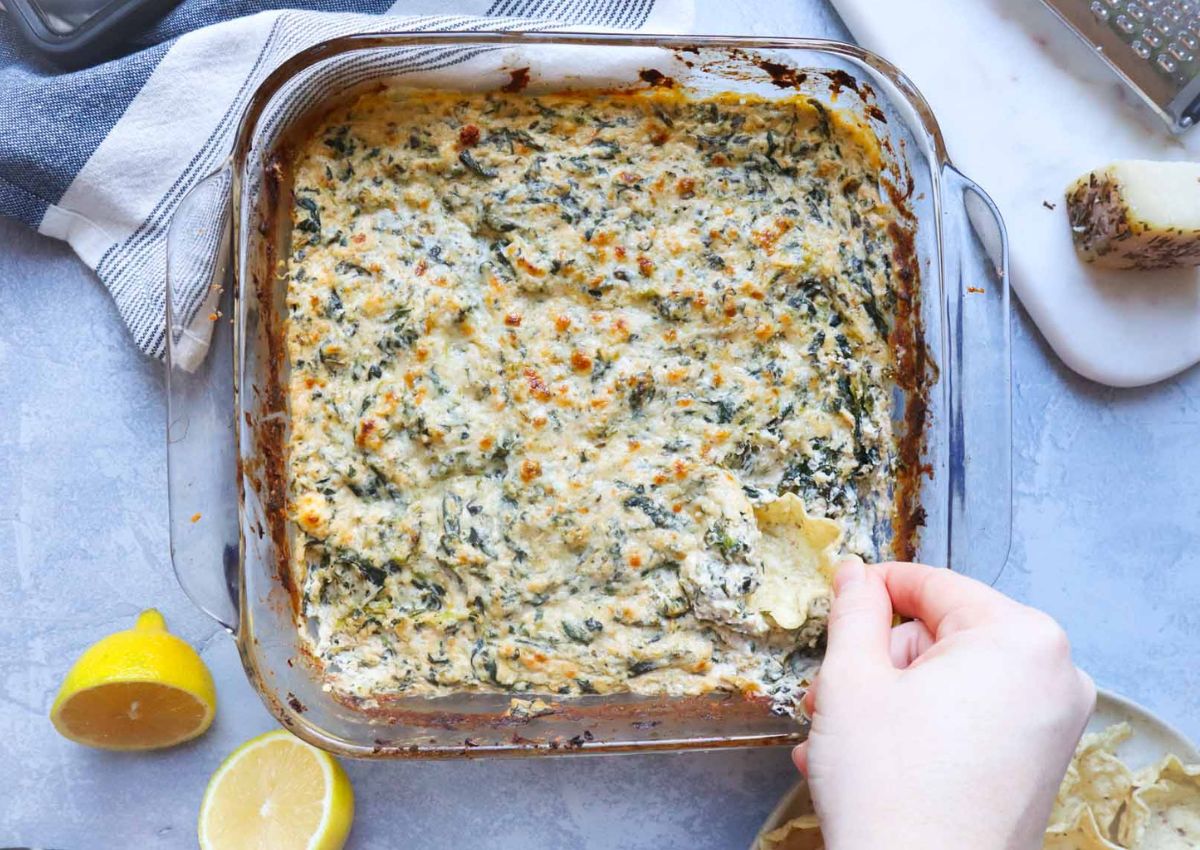 A person dips a chip into a baked spinach and cheese dish in a clear square baking dish.