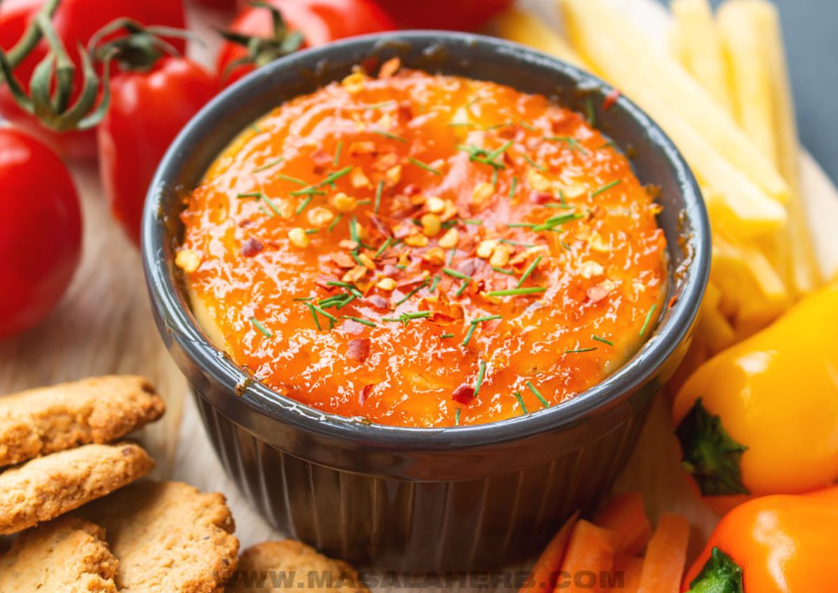 A black bowl filled with baked ricotta is surrounded by bright red tomatoes, yellow bell peppers and thin breadsticks.