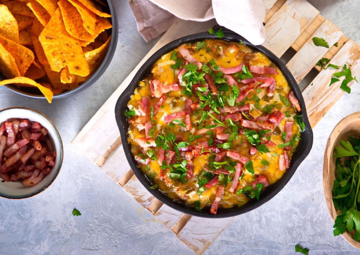 A skillet filled with melted cheese, bacon strips, and chopped green herbs sits on a wooden board.