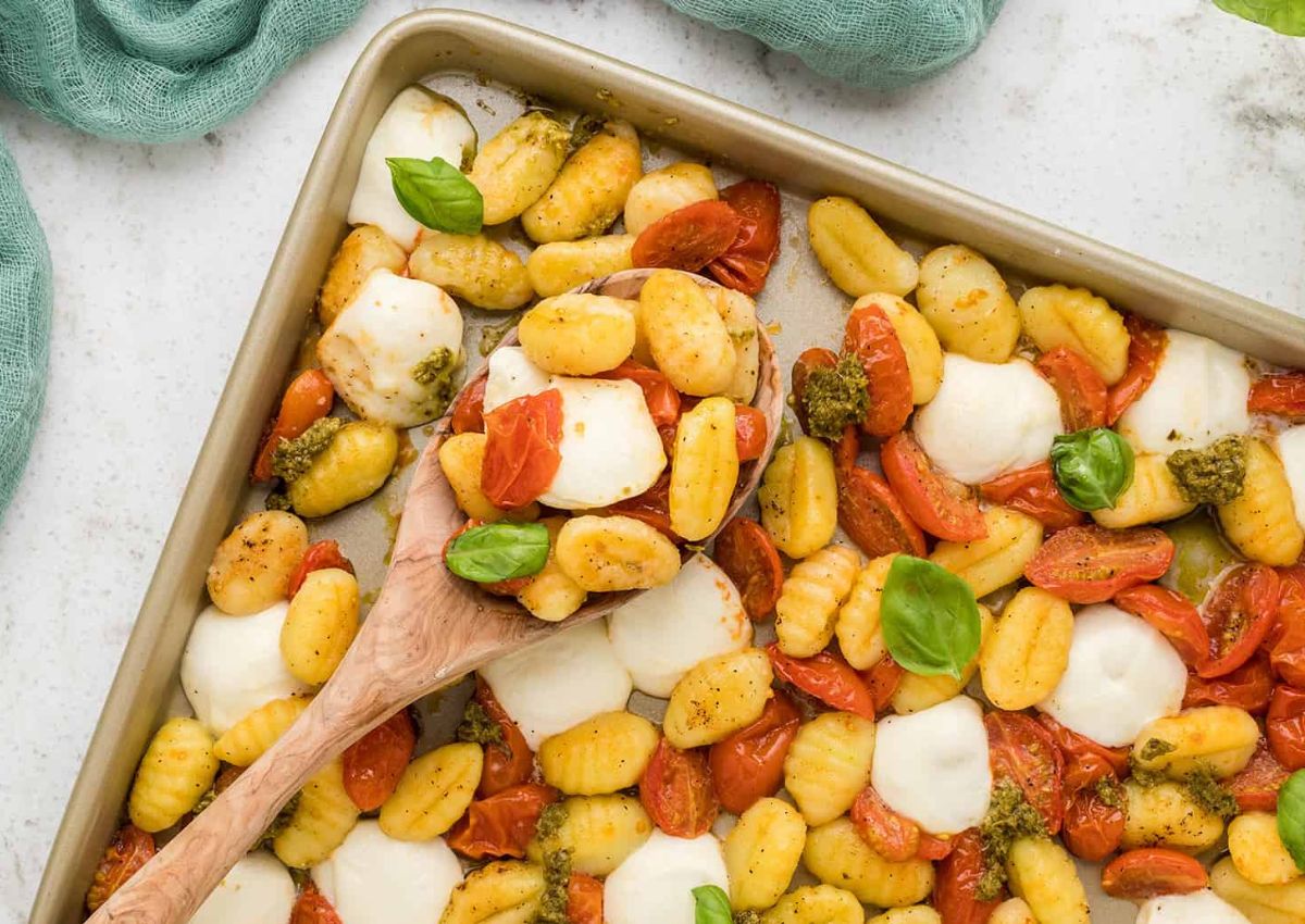 A baking sheet filled with roasted gnocchi, cherry tomatoes, melted mozzarella slices, and dollops of pesto. A wooden spoon is scooping a portion, and fresh basil leaves garnish the dish.