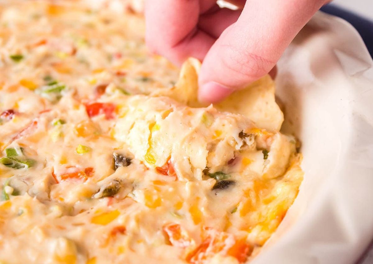 A close-up of a hand dipping a chip into a creamy, cheesy dip with visible pieces of red and yellow bell peppers and green herbs in a dish.