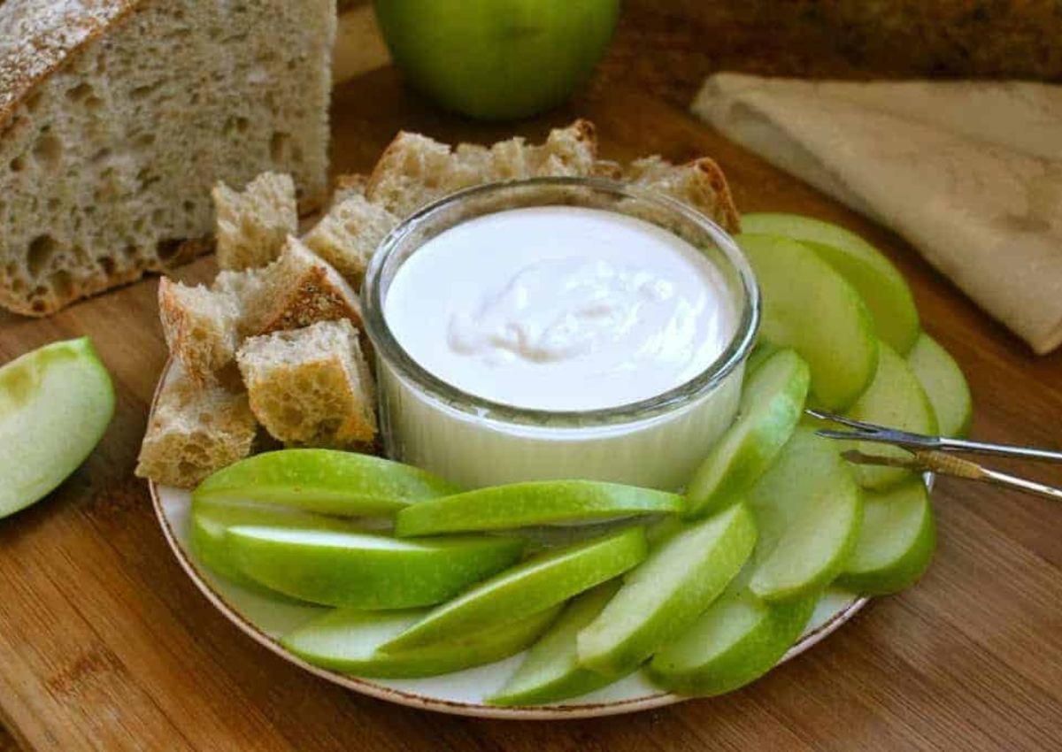 A plate featuring apple slices and chunks of bread surrounds a small bowl of creamy goat cheese dip. 