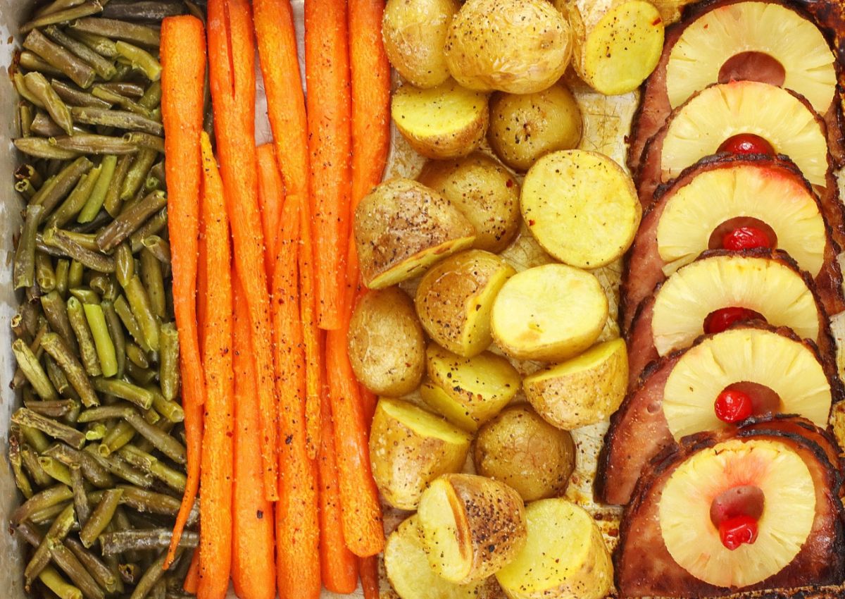 A tray of baked ingredients arranged in rows: green beans on the left, peeled carrots next, then sliced roasted potatoes. On the right are slices of ham topped with pineapple rings and cherries.