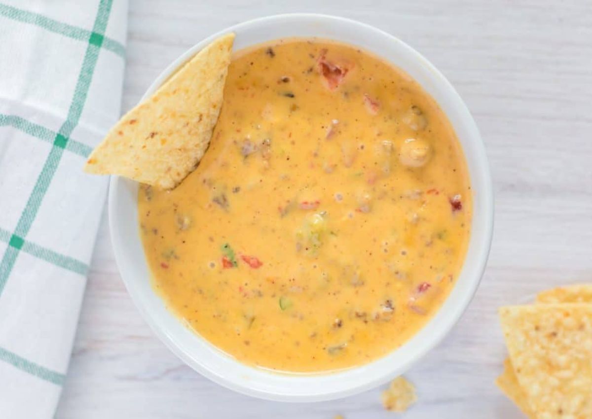 A bowl of cheese dip with visible chunks of vegetables and spices is on a light wooden surface.