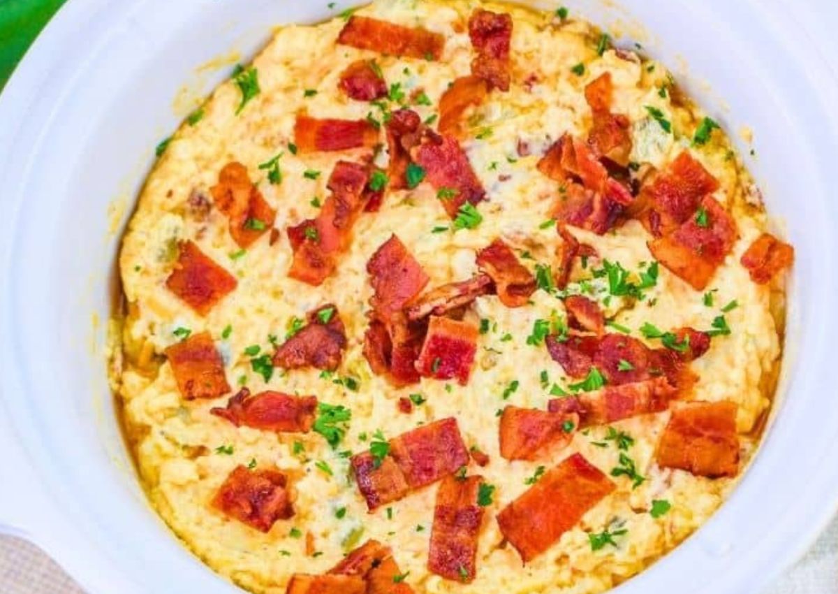 A creamy dish in a white bowl, topped with chopped cooked bacon and garnished with small green herbs.