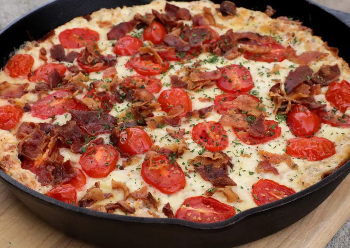 A skillet dish featuring a baked mixture topped with cherry tomato halves, crispy bacon pieces, and garnished with chopped herbs.