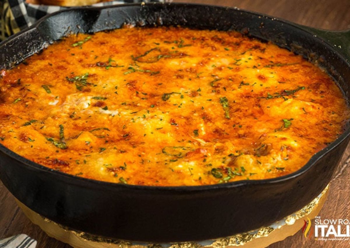 A cast iron skillet filled with baked lasagna dip topped with a golden-brown layer of cheese. Bits of herbs are sprinkled on top. 