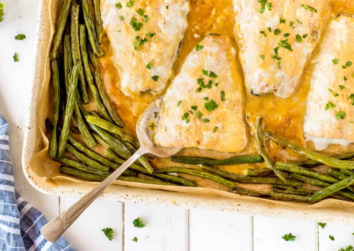 Baked fish fillets topped with herbs in a sauce, surrounded by green beans in a baking dish. A spoon rests on one fillet.