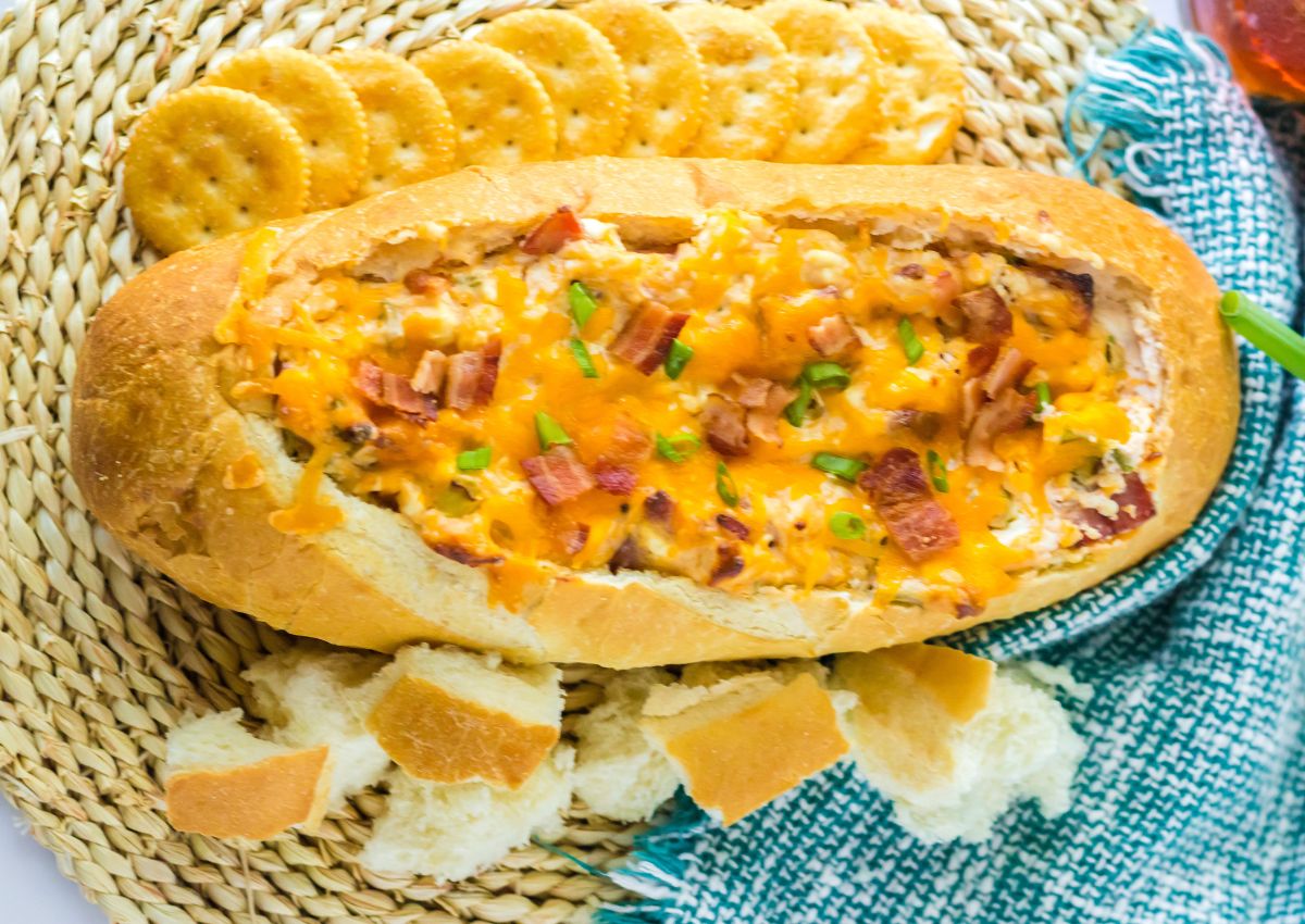 A bread bowl filled with cheesy bacon dip sits on a woven mat, surrounded by round crackers.