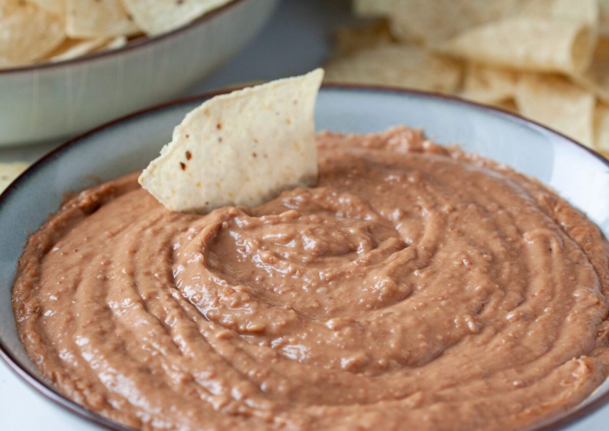A ceramic bowl filled with smooth refried beans, with a single tortilla chip partially submerged in the center.