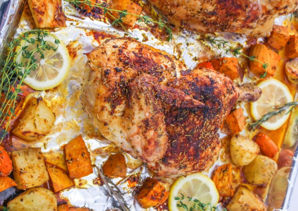 Roasted chicken seasoned with herbs is on a foil-lined tray surrounded by chunks of roasted potatoes, carrots, and lemon slices. Sprigs of fresh thyme are scattered around, adding to the rustic presentation.