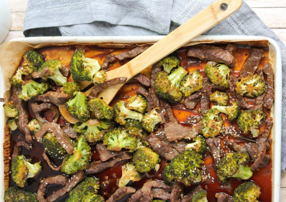 A baking tray filled with cooked beef strips and broccoli pieces, garnished with sesame seeds. A wooden spatula rests on top.