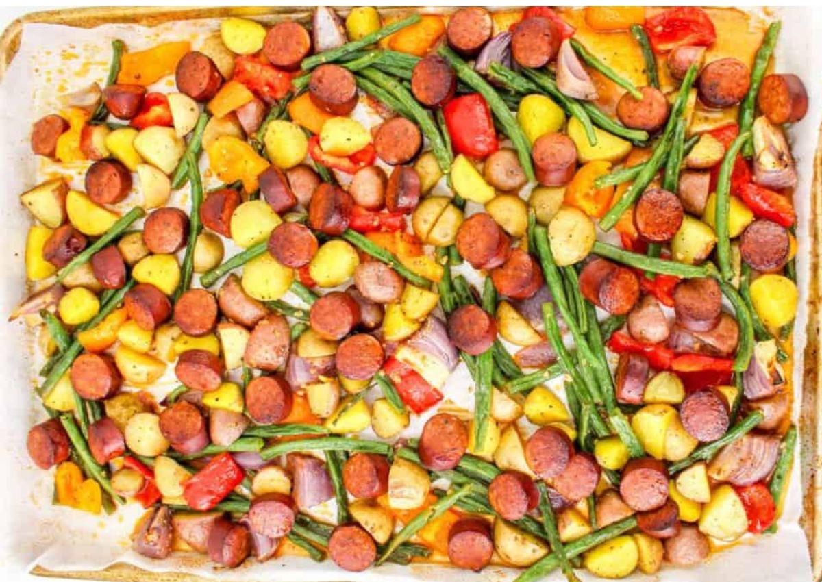 A baked sheet pan meal featuring sliced sausage, halved baby potatoes, green beans, and chunks of red and yellow bell peppers on parchment paper.