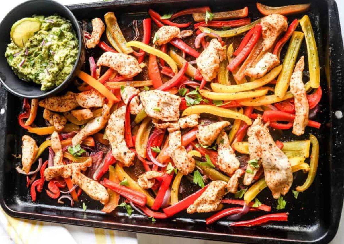 A baking sheet filled with cooked chicken strips, and red, yellow, and orange bell peppers. A small black bowl of guacamole is placed on the top left corner.