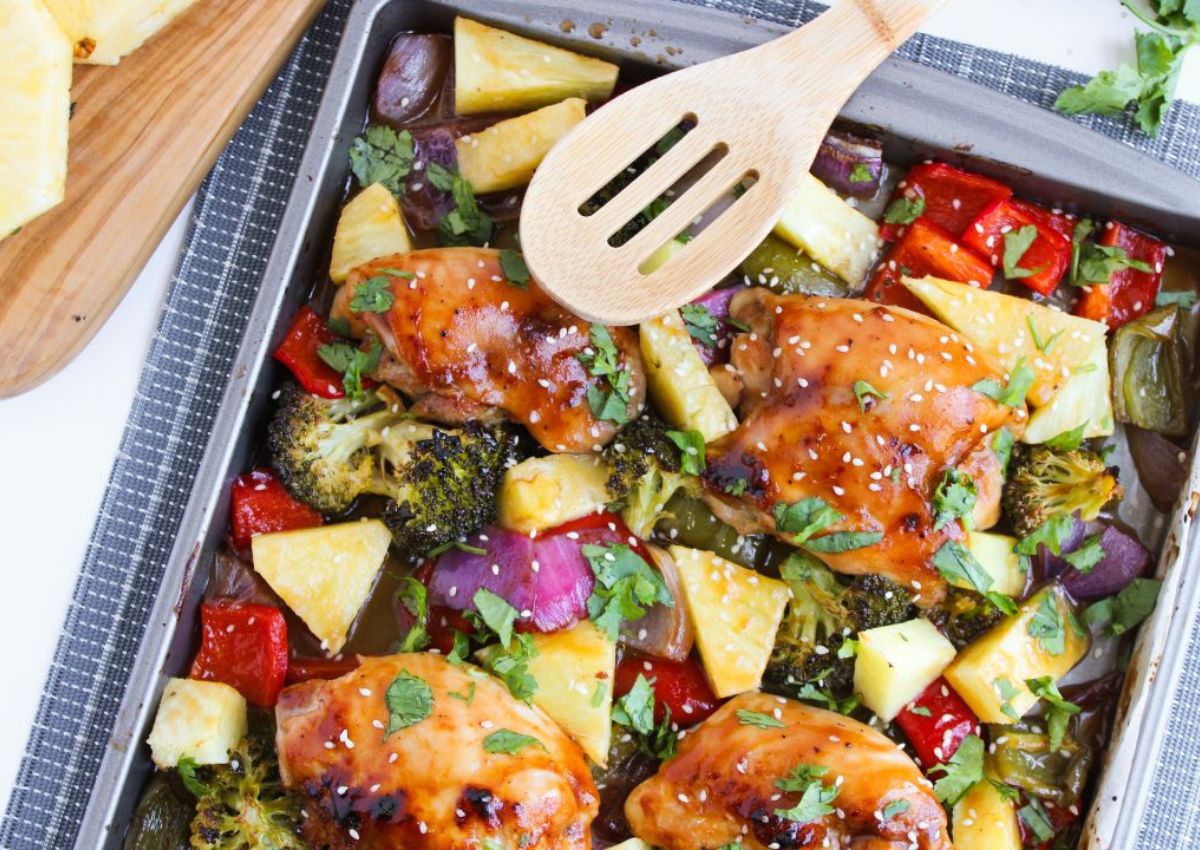 A baking tray contains cooked chicken pieces, pineapple chunks, broccoli, red and green bell peppers, and red onions, garnished with herbs.