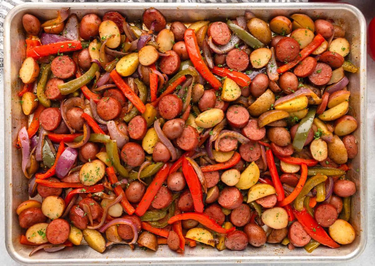 A baking tray filled with a mix of sliced sausages, red and green bell peppers, red onions, and halved small potatoes.