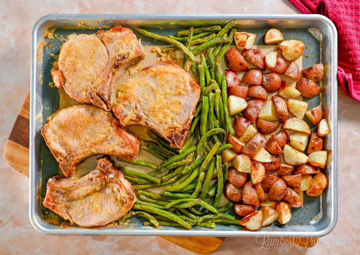 A baking sheet with cooked pork chops, roasted red potatoes, and green beans. The pork chops are seasoned, and the vegetables appear lightly browned.