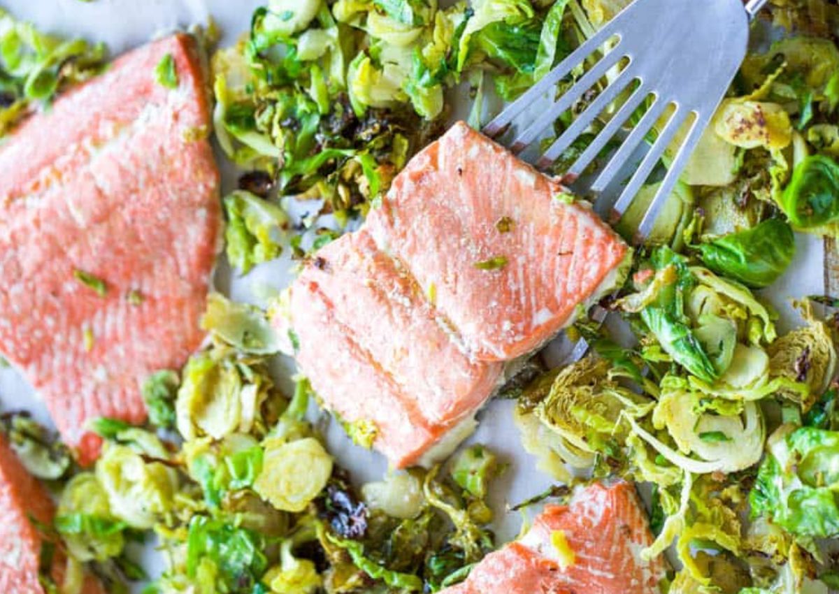 A cooked salmon fillet being lifted by a spatula is surrounded by roasted Brussels sprouts on a pan.