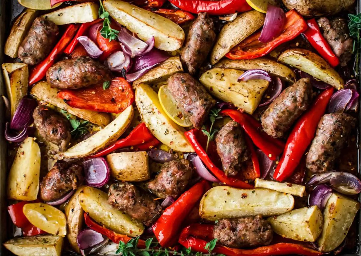 A tray of roasted sausage pieces, potato wedges, red bell pepper slices, and red onion chunks, garnished with fresh herbs.