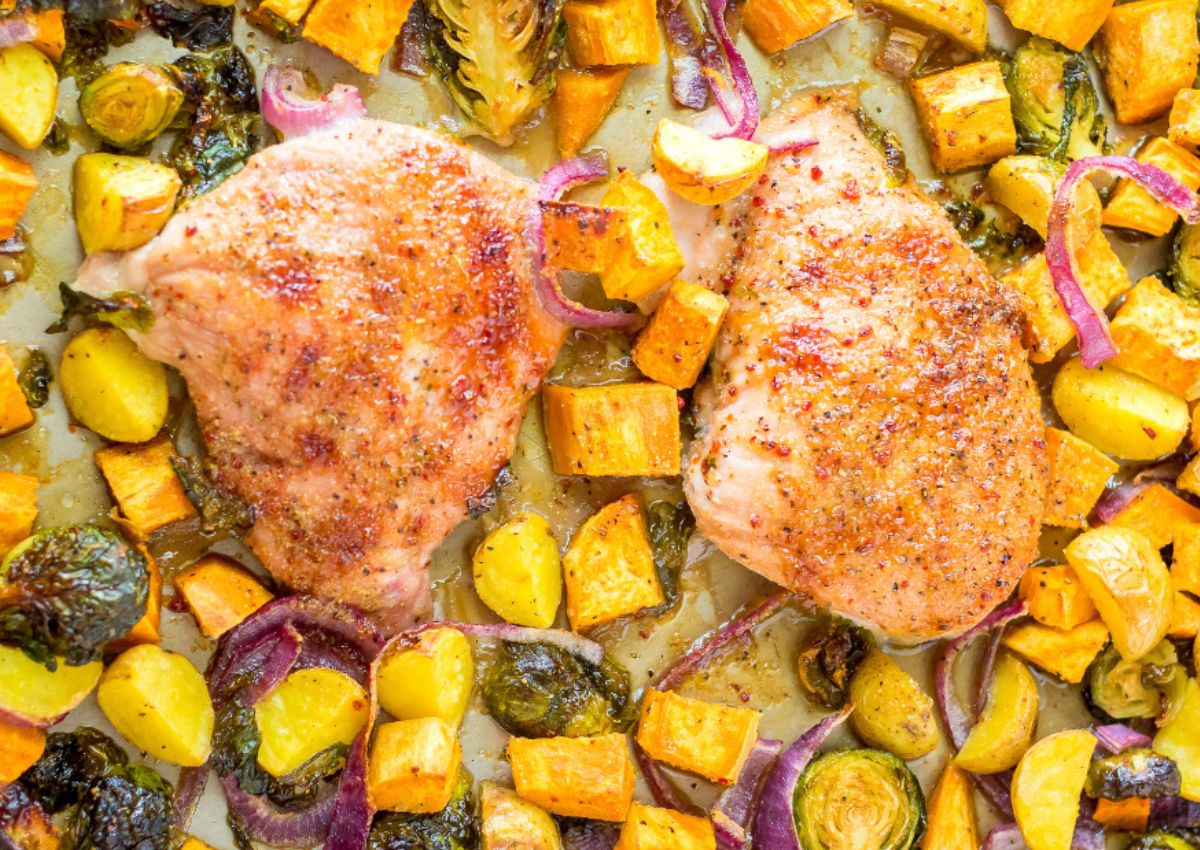 A baked dish featuring two seasoned chicken breasts surrounded by roasted brussels sprouts, sweet potatoes.