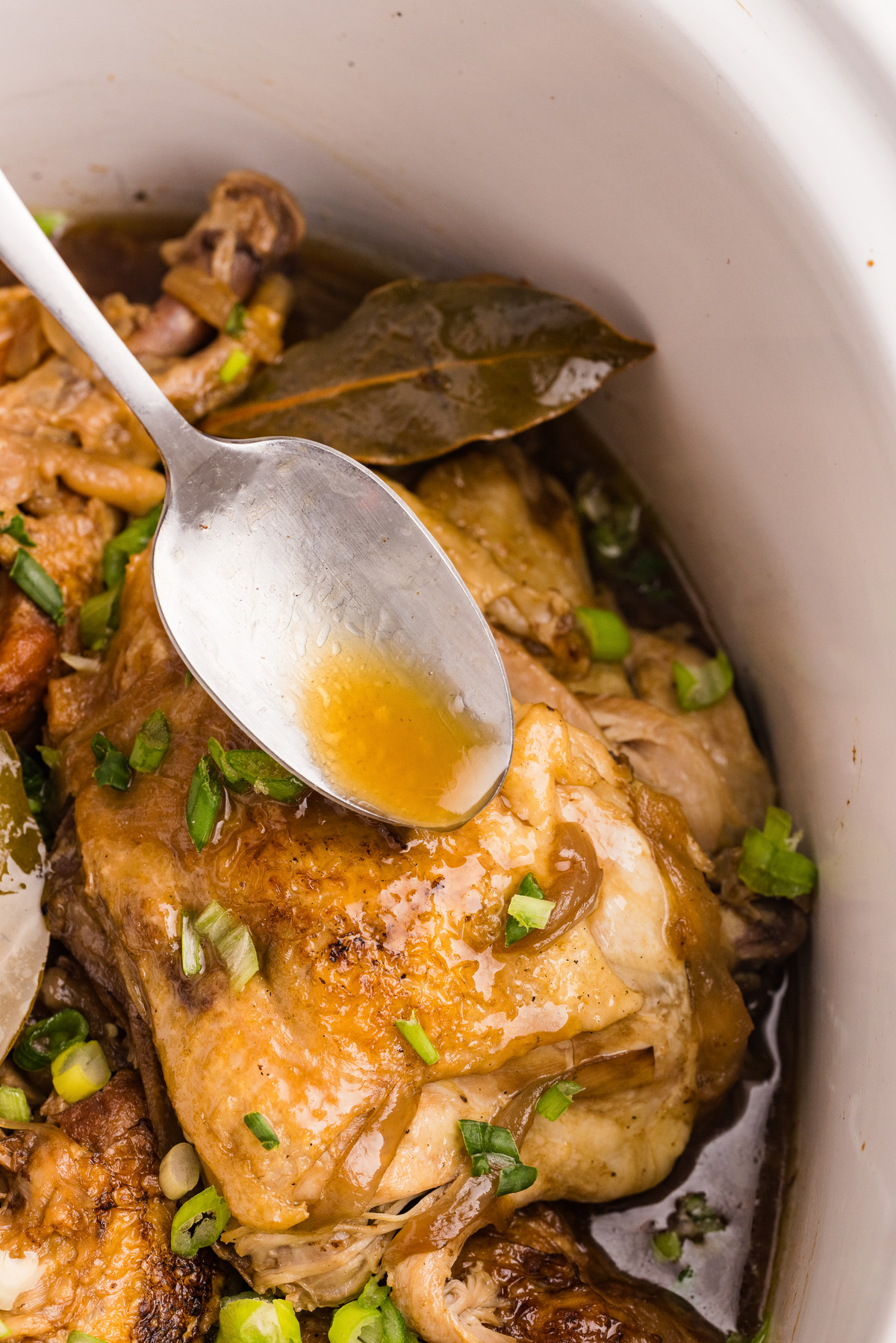 Chicken pieces in a slow cooker with a spoon full of adobo juice.