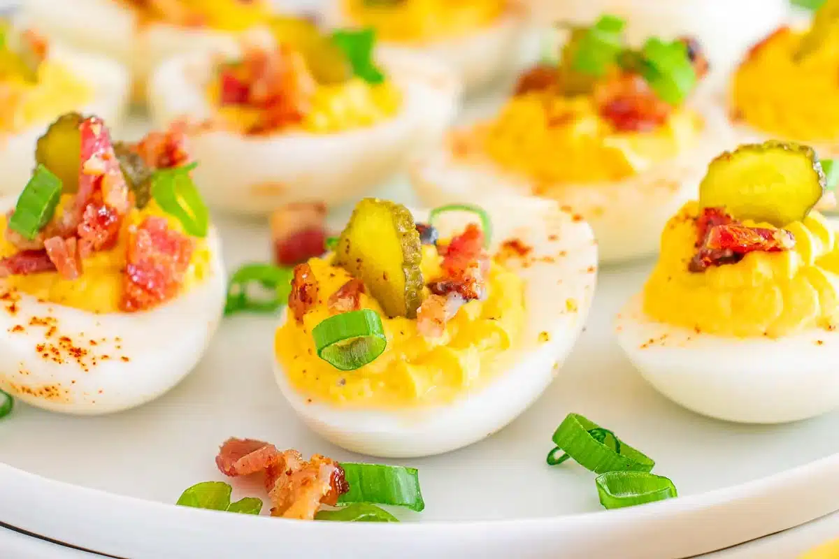Deviled eggs topped with pickles, green onions, and bacon on a white plate.
