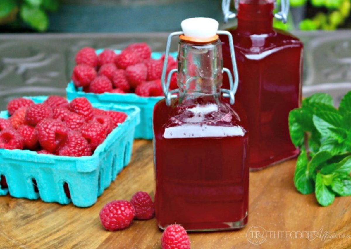 Bottles of raspberry syrup on a counter with baskets of fresh raspberries.