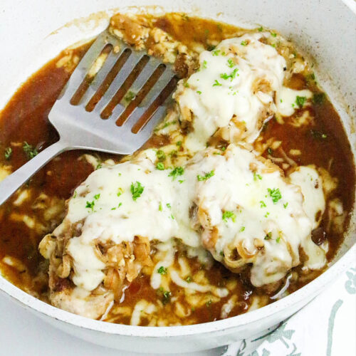 French onion pork chops in a white skillet topped with melted cheese.