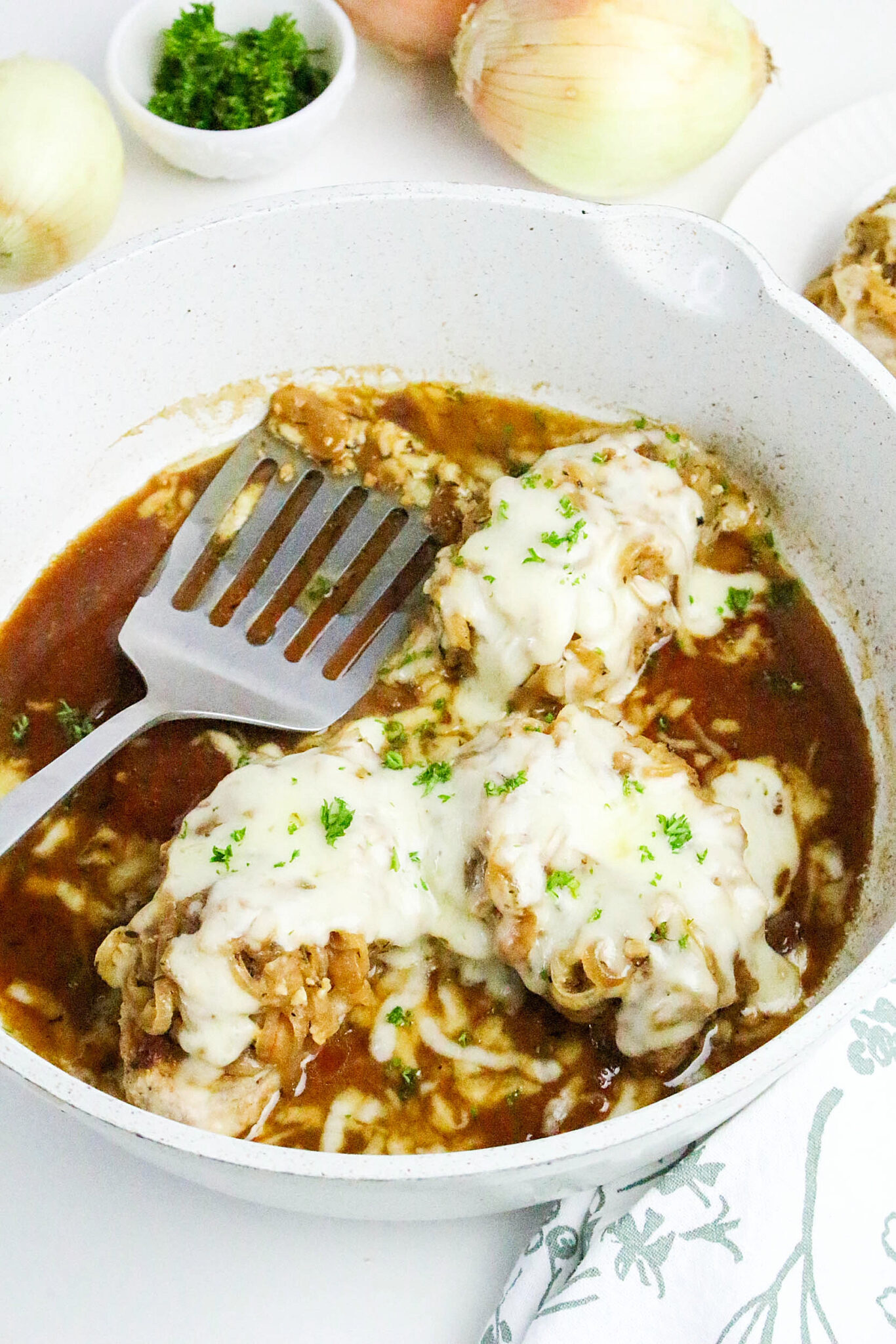 White skillet with a silver spatula with pork chops covered with onions and melted cheese.