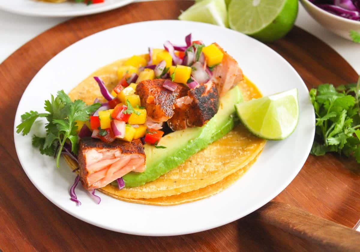 Blackened salmon tacos with mango salsa, red cabbage, and avocado slices on a plate.