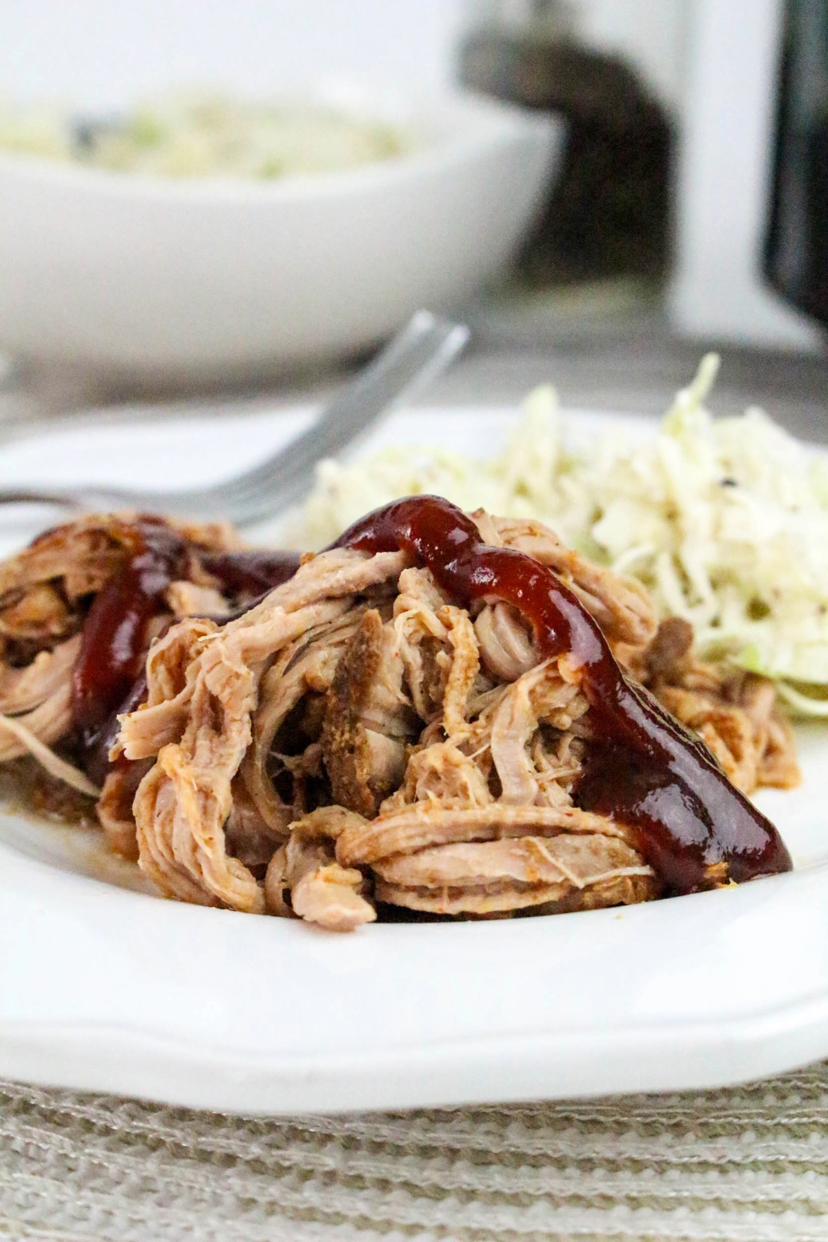 A white plate with shredded pulled pork tenderloin topped with BBQ sauce with coleslaw on the side.