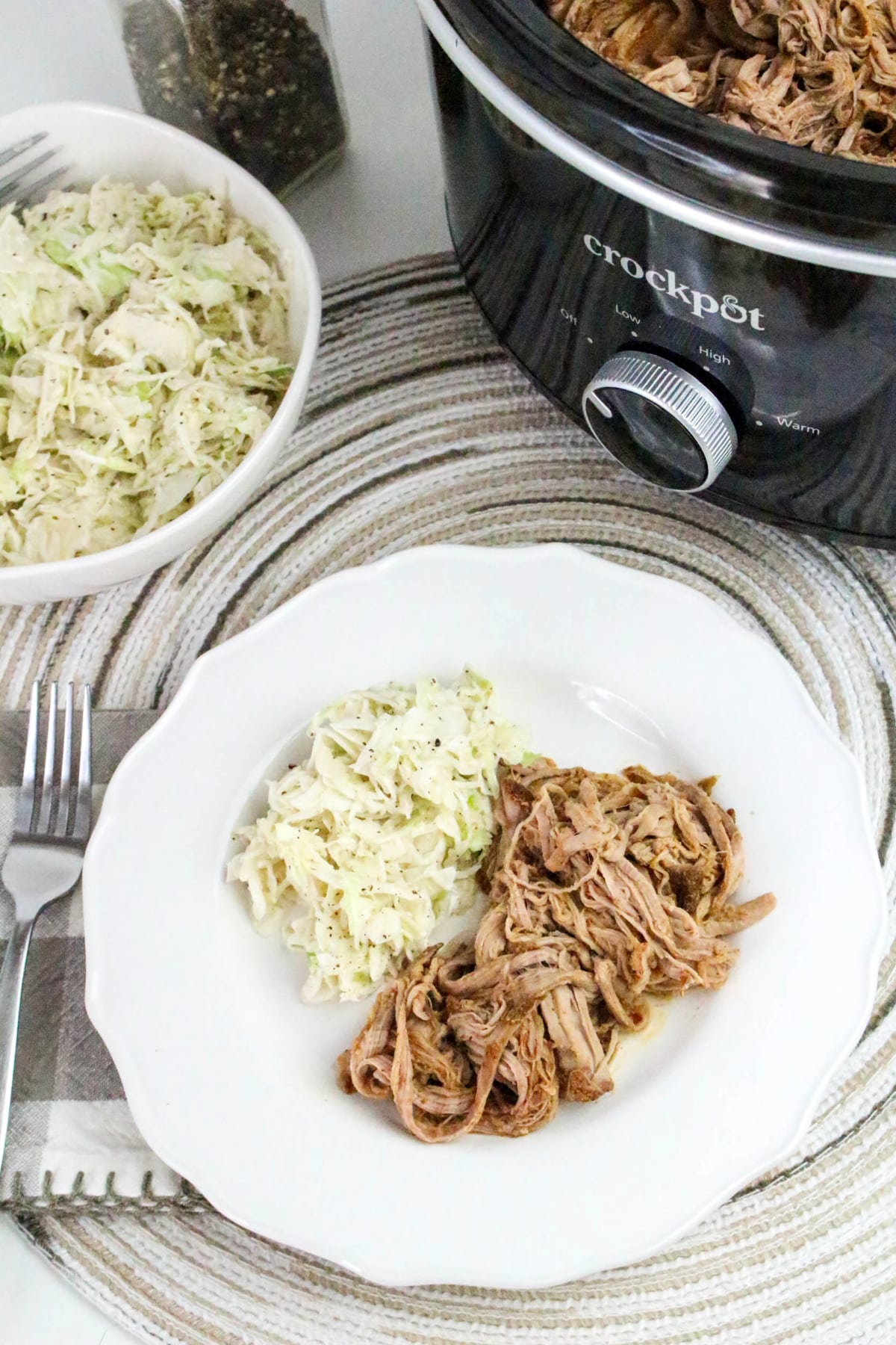 Top view of a dinner plate with shredded pork and coleslaw with a black slow cooker on the side.