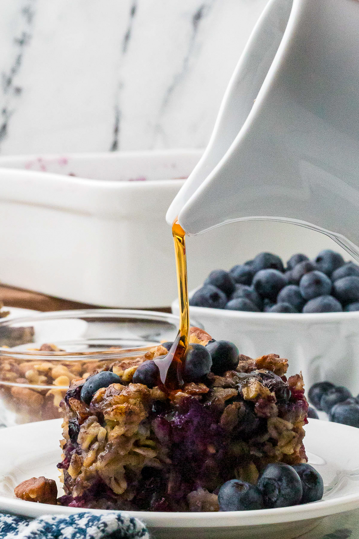 A white small pitcher with maple syrup pouring over slice of baked oatmeal with blueberries.